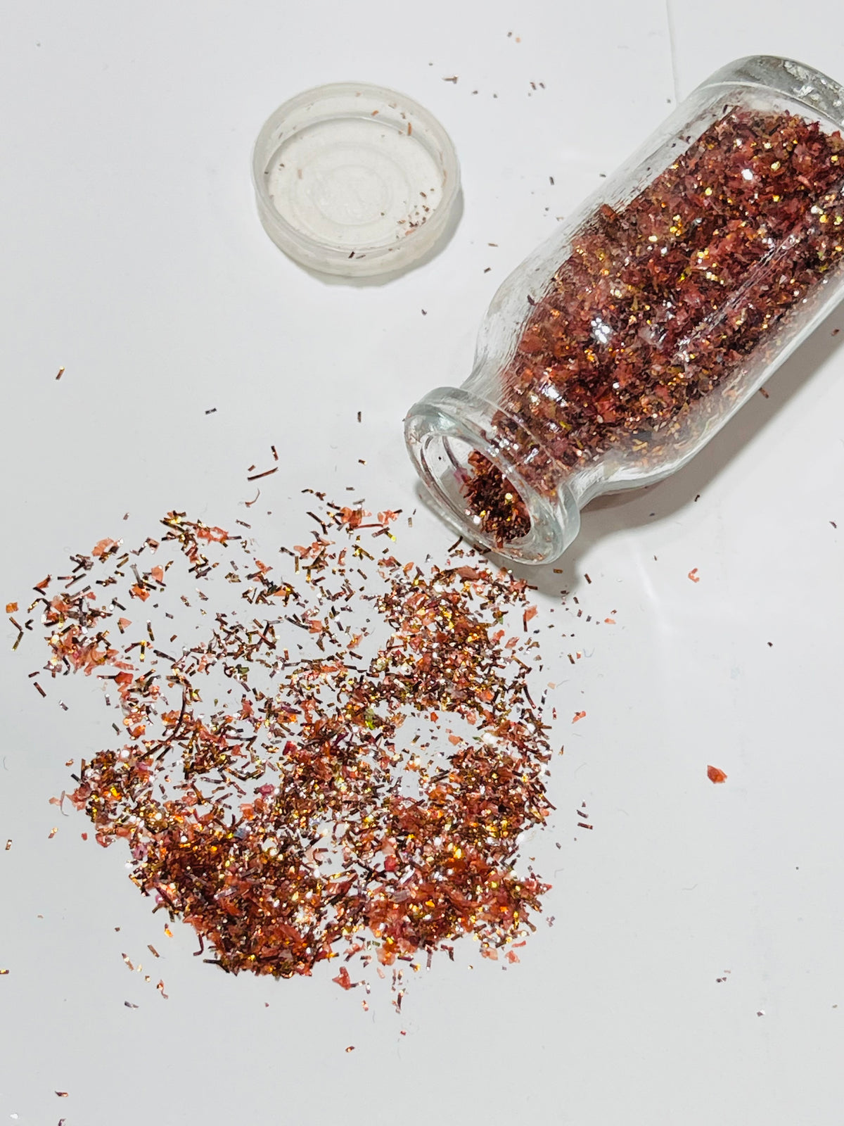 Red glitter flakes spilled from a clear glass bottle onto a white surface, with the bottle lid placed nearby.
