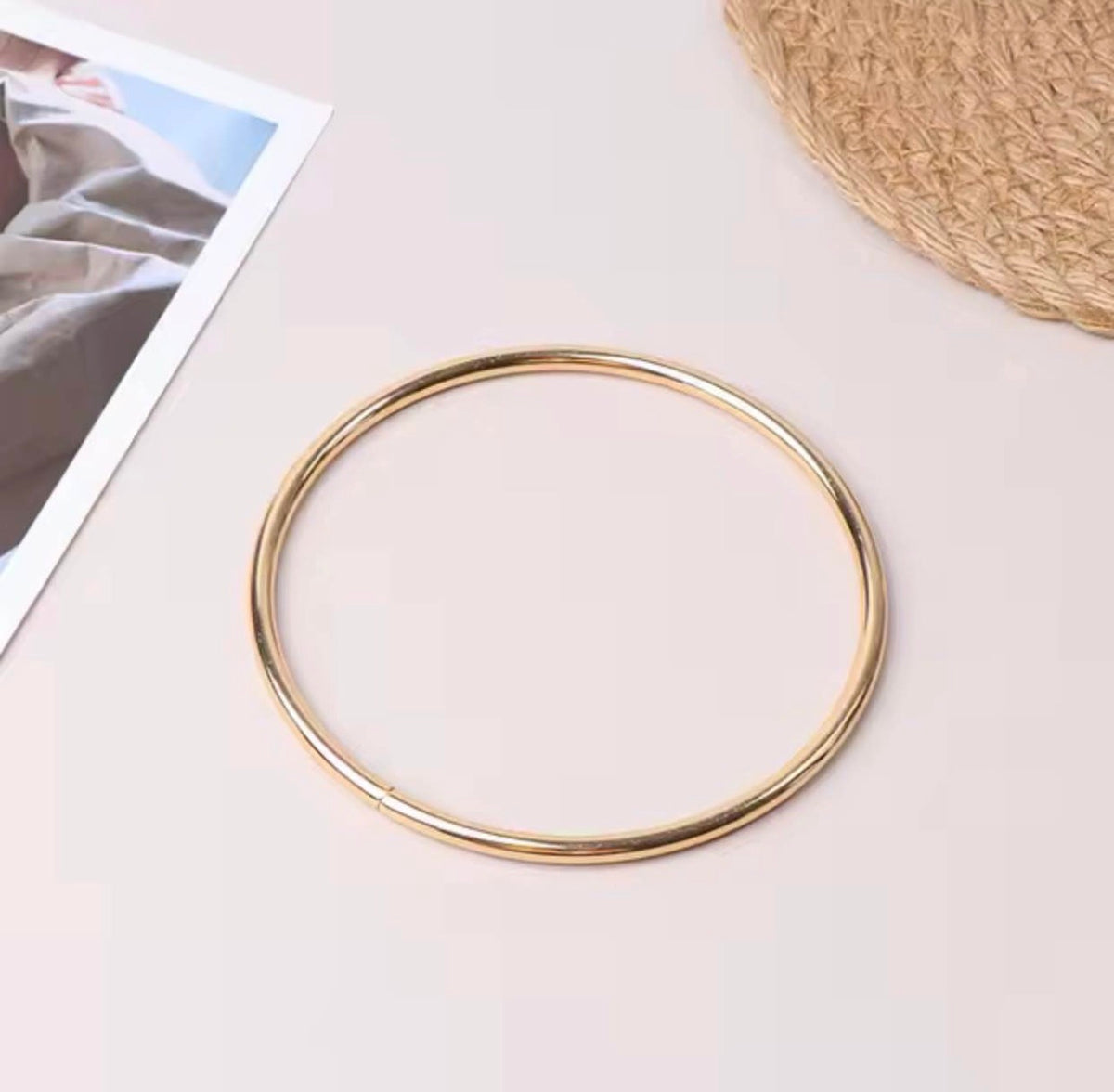 A thin, polished gold metal bangle bracelet displayed on a light pink background with styled flat-lay props, including a printed photo card and a woven straw hat corner.