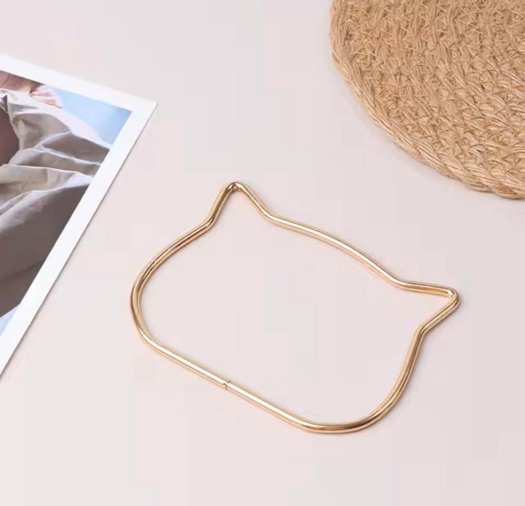A gold metal cat-shaped frame or hoop laid on a light beige surface, with part of a printed photo and a woven straw hat visible as styled props.