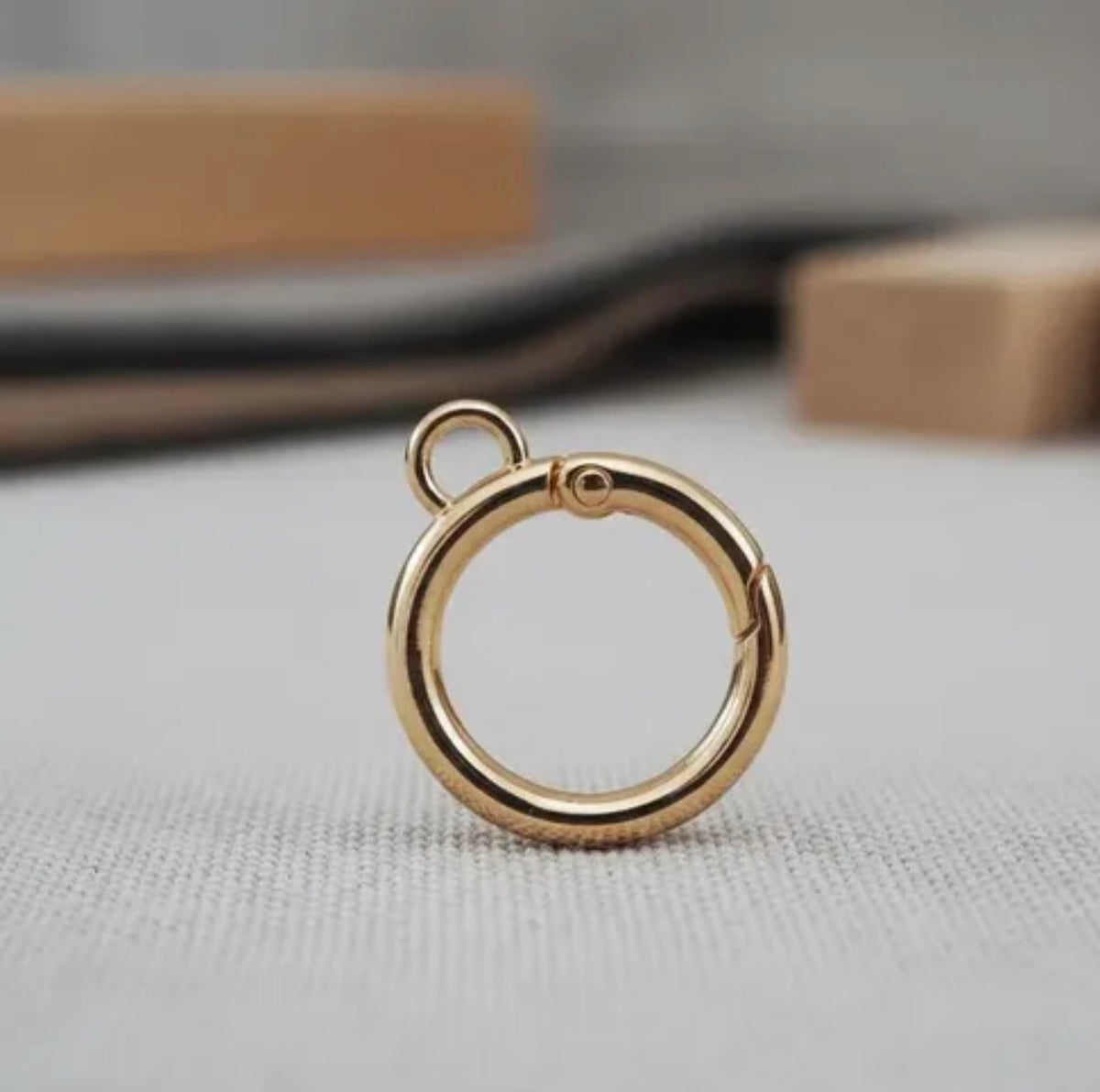 A polished gold-tone metal jump ring with a small attached loop, resting on a light gray textured surface with a blurred background.