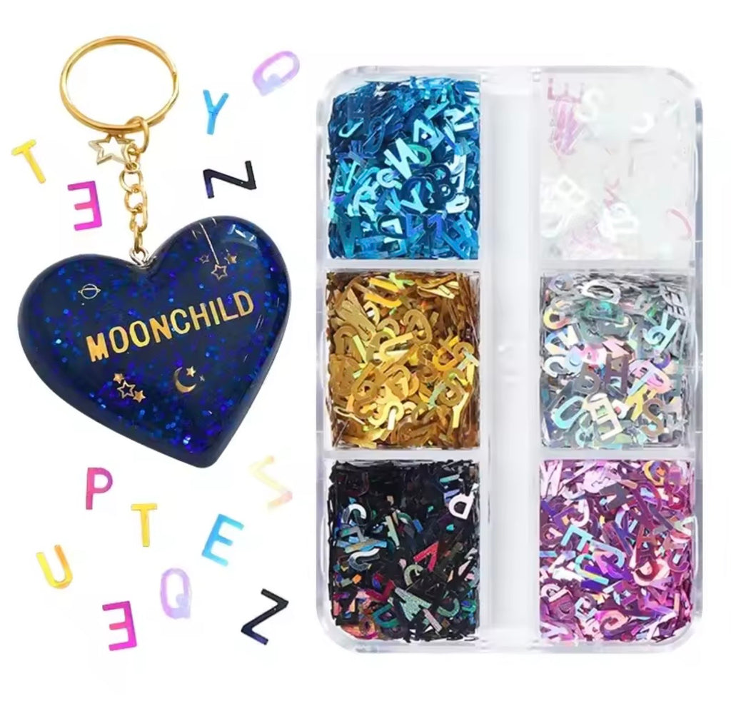 A heart-shaped keychain with a gold ring and chain, featuring a dark blue glittery face with the text 'MOONCHILD' and small celestial motifs, shown next to a clear plastic organizer filled with colorful letter-shaped glitter in multiple colors.