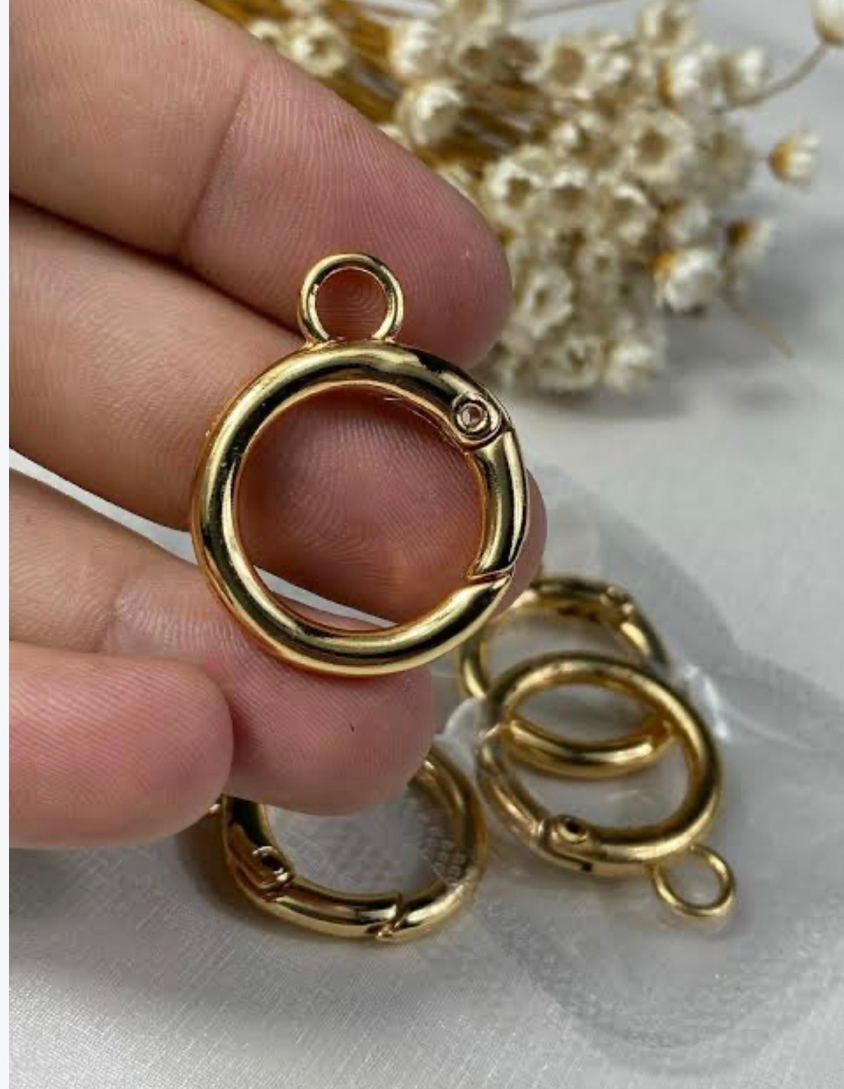 A hand holding a polished gold-plated jump ring clasp, with additional matching jump rings resting on a white surface in the background.