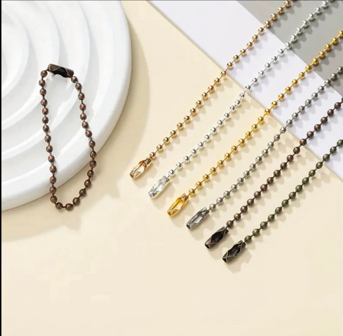 A set of beaded keychains or phone straps in multiple metallic finishes, including dark bronze, silver, gold, and black, arranged on a beige background with a white ceramic dish.