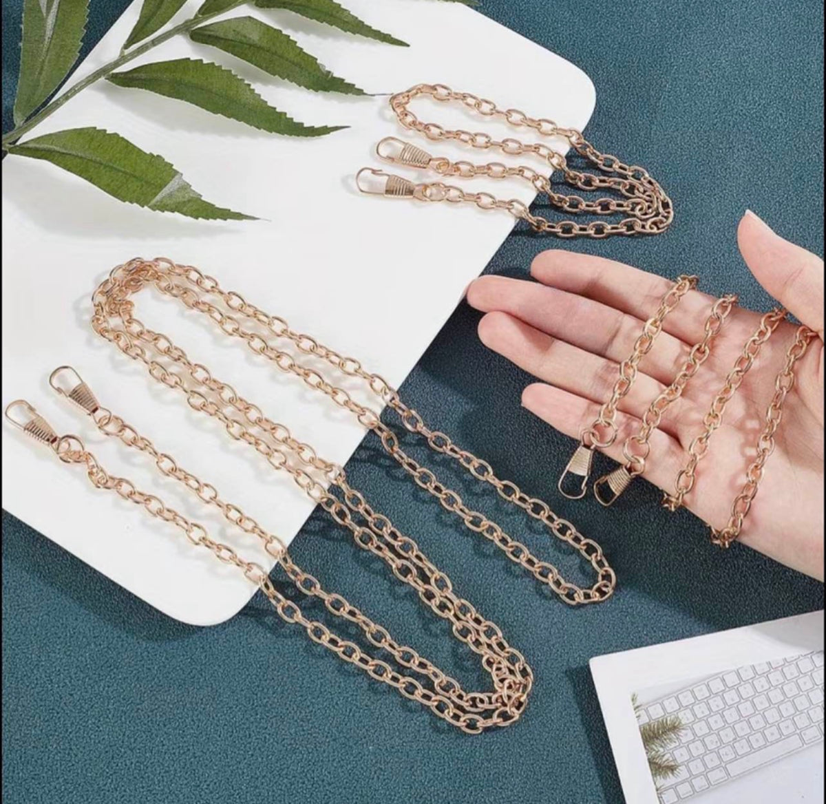 Rose gold chain straps with lobster clasp ends, shown draped on a white card and held in a hand against a teal textured background with a green leaf and keyboard prop.