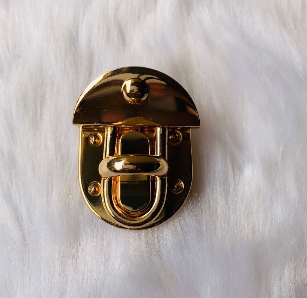 A polished gold metal latch or clasp with a U-shaped bar and rivet details, photographed from above on a white faux fur background.