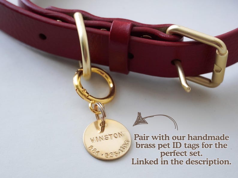 A close-up of a burgundy leather pet collar with gold-tone hardware and a round engraved brass tag hanging from a ring. Text on the image suggests pairing with handmade brass pet ID tags.
