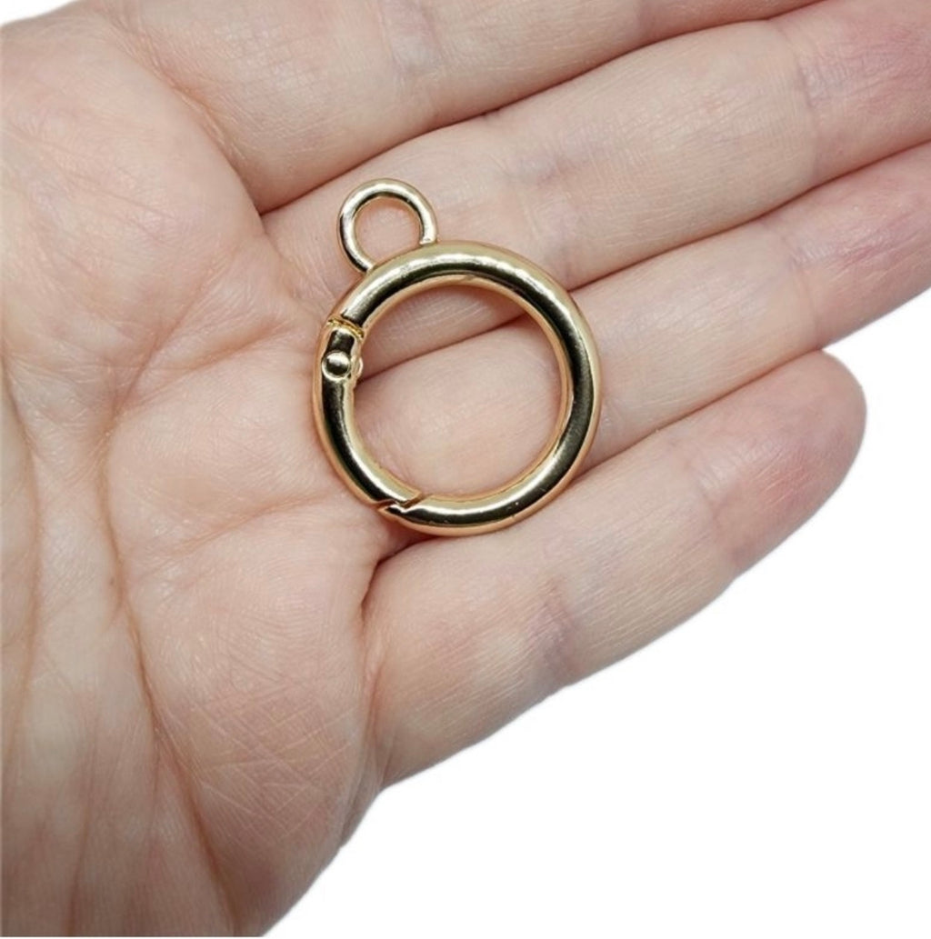 A polished gold-toned split ring clasp with a small top loop, shown resting on an open hand against a white background.