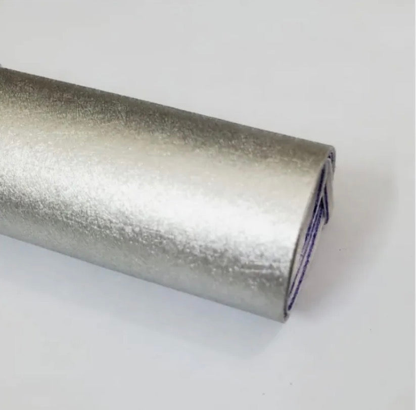 A close-up of a rolled metallic silver material, likely aluminum foil or a silver adhesive film, showing its textured reflective surface and purple backing layer.