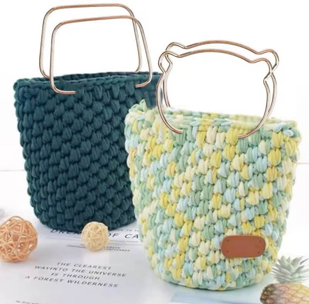 Two chunky knit bucket handbags with metal cat-ear style handles, displayed on a white background with decorative props. The bag in the back is dark teal, and the bag in the front is a variegated light green, yellow, and white pattern with a small tan leather patch.