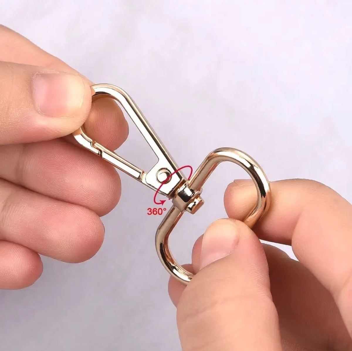 A close-up of a person's hands holding a gold-toned metal swivel clasp with a split ring, showing the 360-degree rotation feature against a light background.