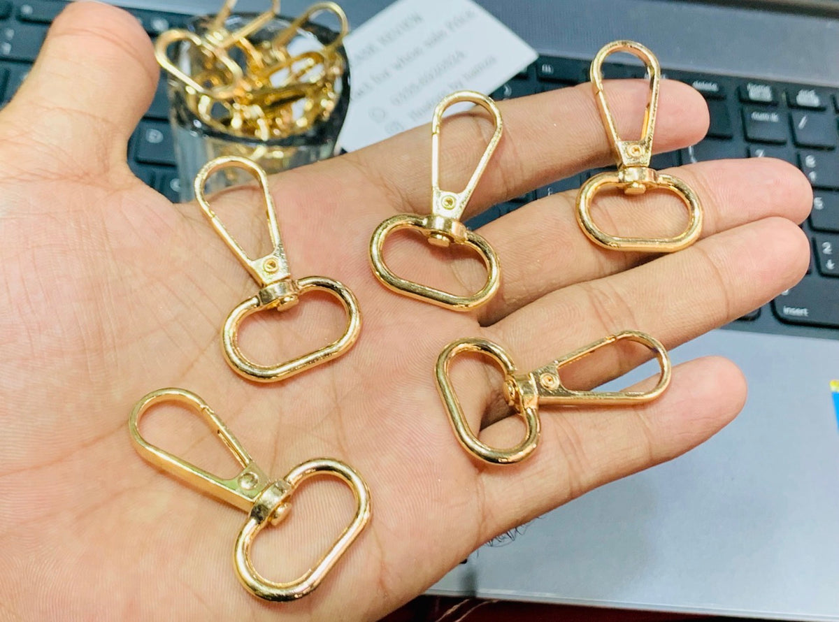 Five gold-colored metal swivel snap hooks held in a person's hand, with a keyboard and a small container of similar hooks in the background.