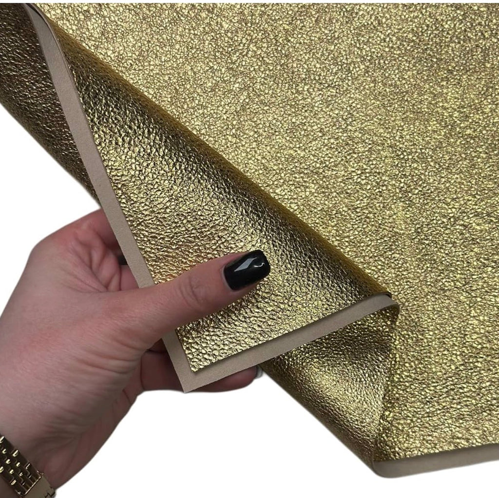 A hand with black nail polish holds a corner of a textured gold adhesive film or sticker, showing its peel-and-stick backing against a plain white background.