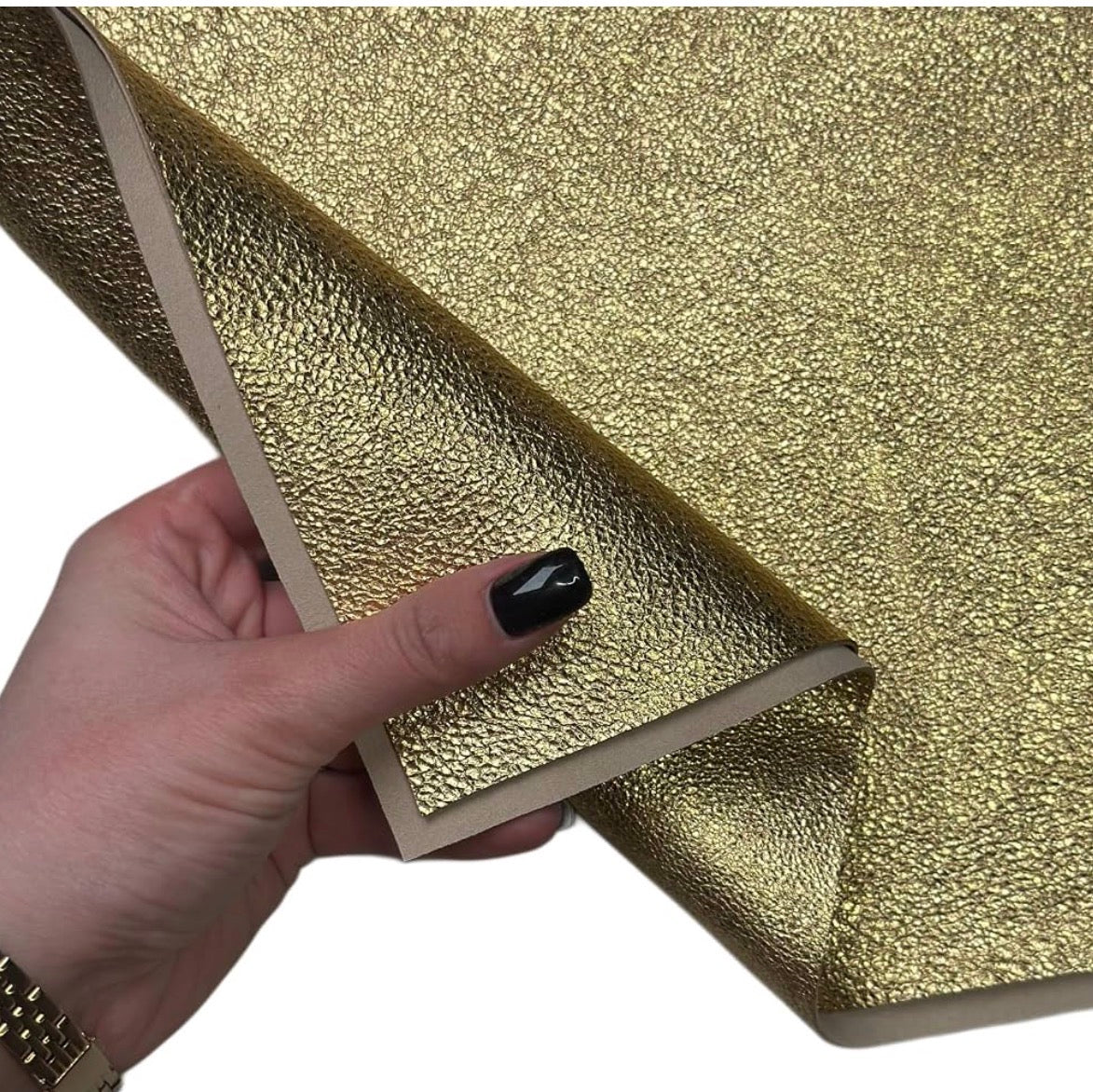 A hand with black nail polish holds a corner of a textured gold adhesive film or sticker, showing its peel-and-stick backing against a plain white background.