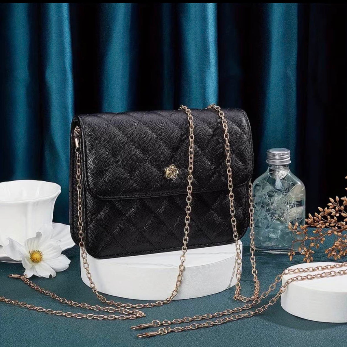A black quilted leather shoulder bag with a gold-tone chain strap and a central gold emblem, displayed on a white pedestal against a dark teal curtain backdrop.