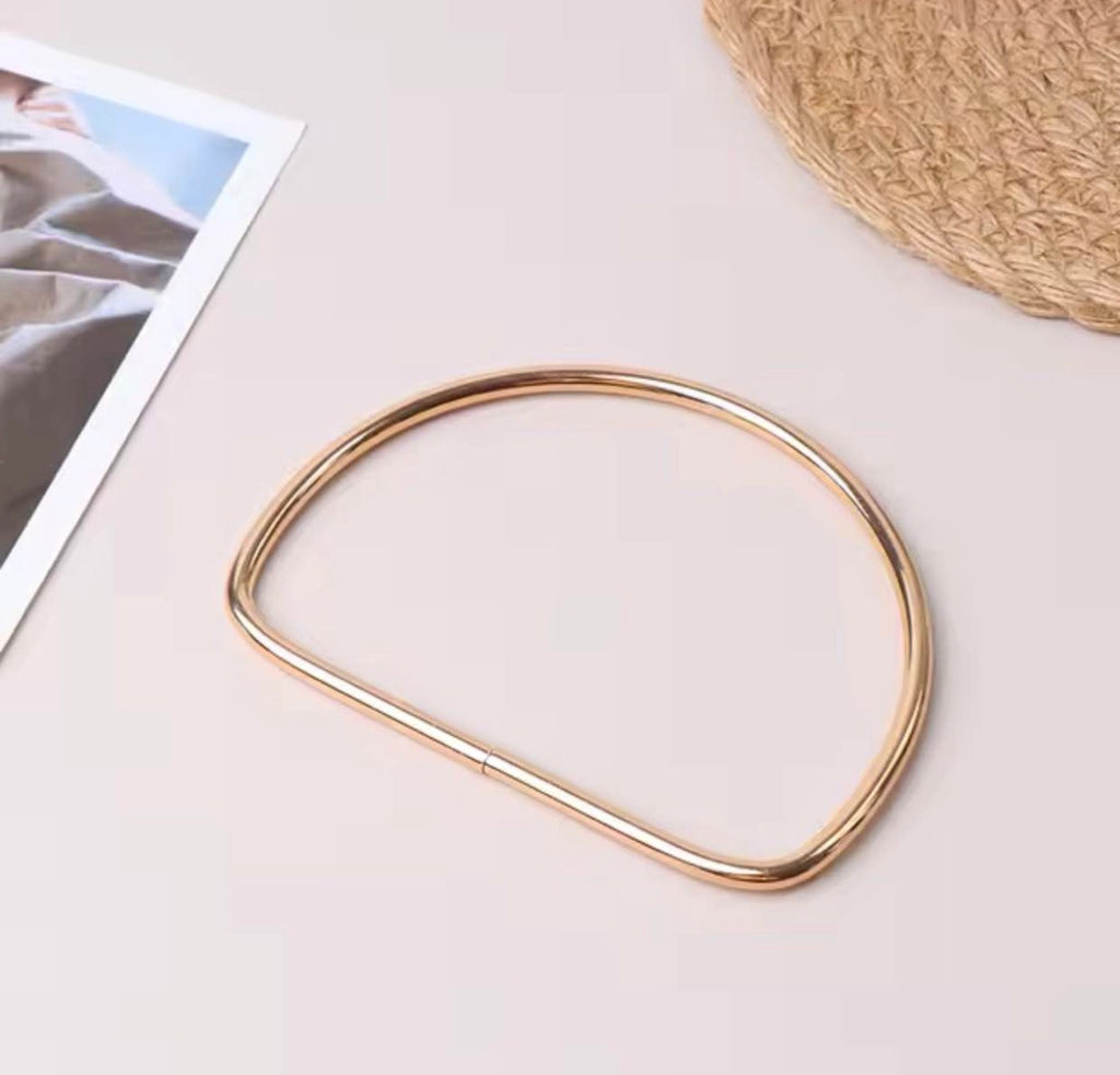 A polished rose gold metal frame or bezel component, shown on a light beige background with styled flat-lay props including a corner of a printed photo and a woven straw hat.