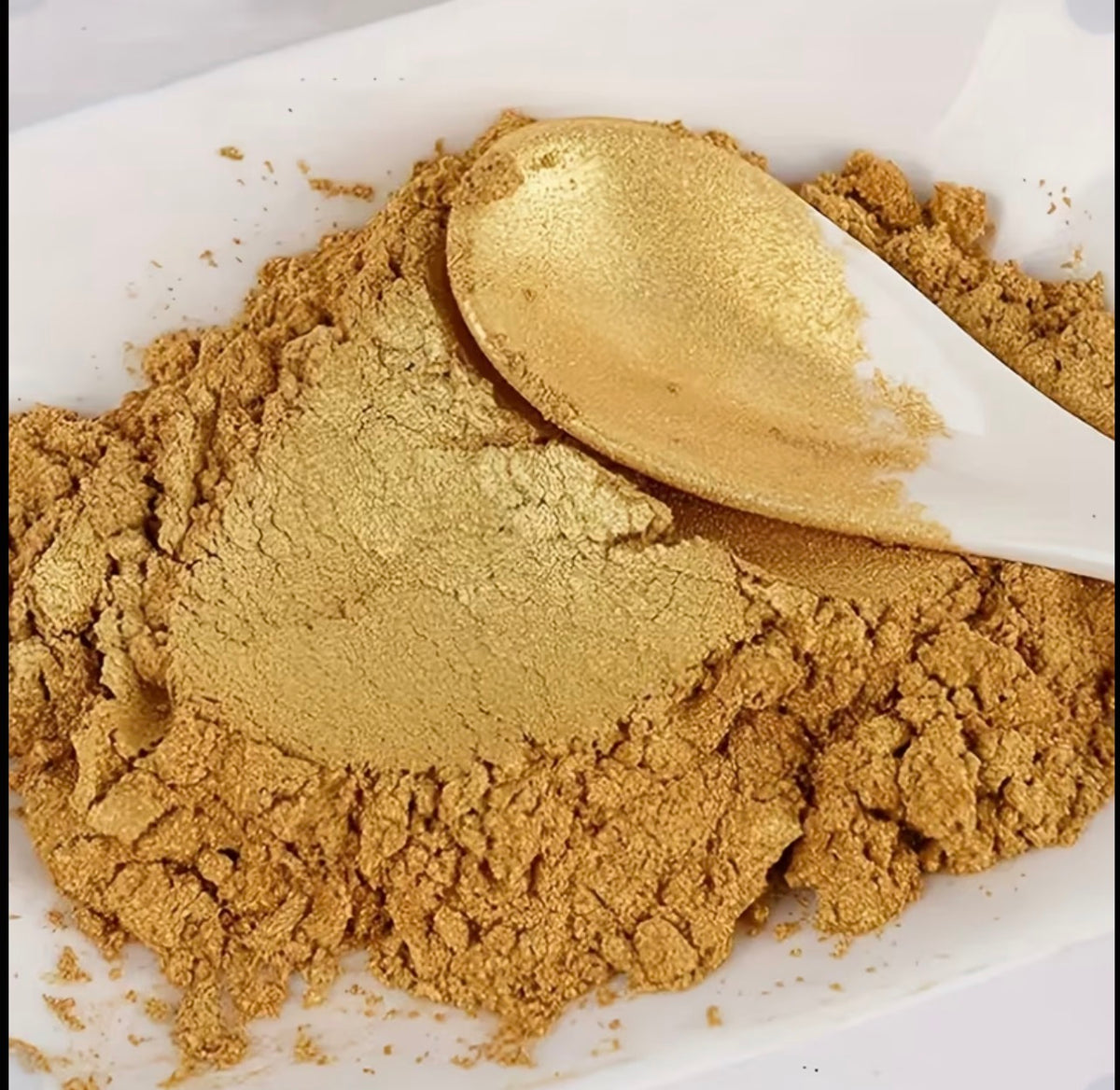 A close-up of a fine golden-brown powder piled in a white dish, with a white spoon resting on top and more powder scattered around.