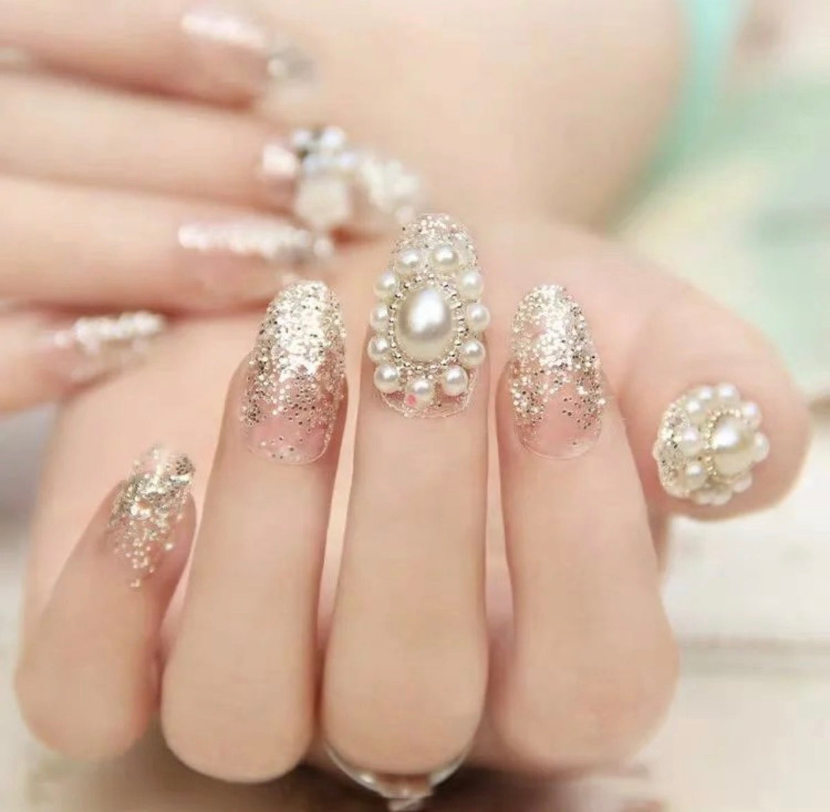 A close-up of a hand showcasing elegant, long press-on nails decorated with glitter, faux pearls, and floral nail art.