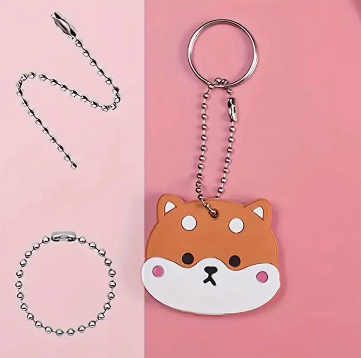 A cute dog-face keychain with a silver ball chain and key ring, shown on a pink background with the chain and ring separated on the left side. The keychain charm is orange-brown and white with pink cheeks and a black nose.