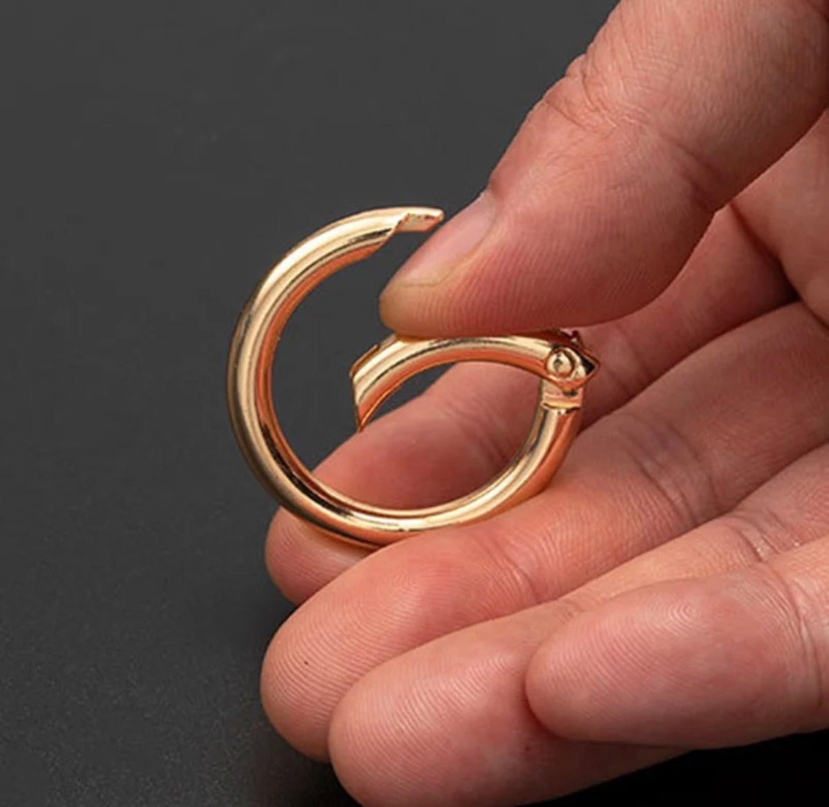 A close-up of a hand holding a small polished gold-colored metal ring or clasp against a dark gray background.