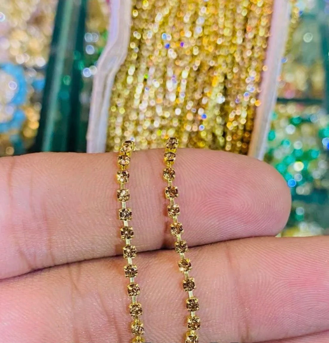 A close-up of a hand holding a delicate gold chain necklace with small, round, textured beads. The background shows a blurred display of other colorful strands of beads.